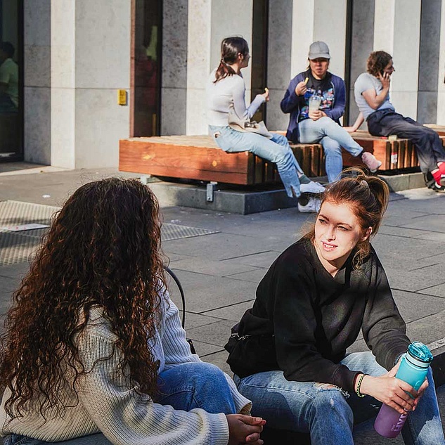 Mehre Studierende sitzen vor dem Grimm-Zentrum der HU und unterhalten sich, telefonieren oder lesen auf dem Handy. Mehre Studierende sitzen vor dem Grimm-Zentrum der HU und unterhalten sich, telefonieren oder lesen auf dem Handy.